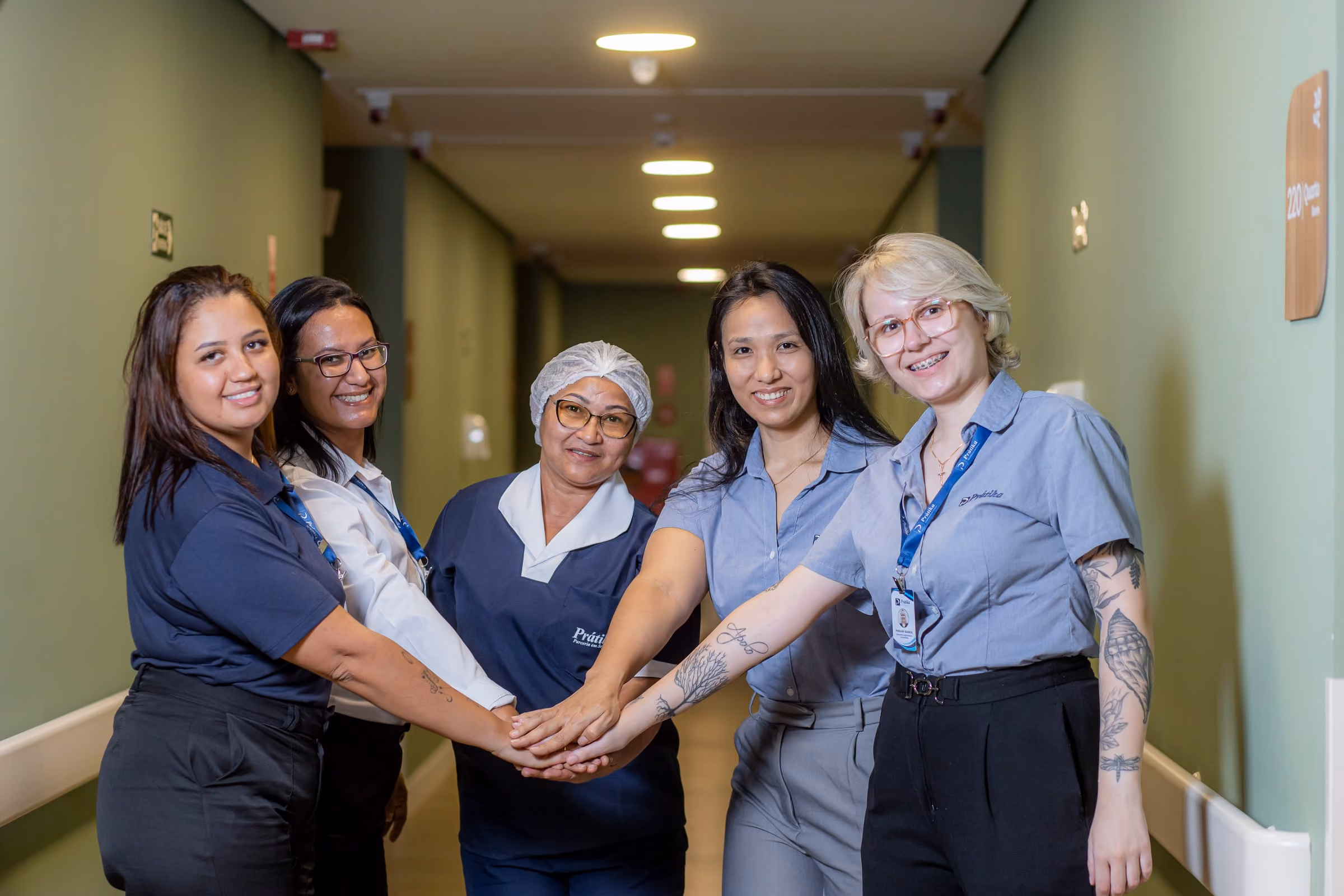 Equipe da Prátika, de limpeza, supervisão e gestão, sorri com as mãos juntas num corredor de hospital